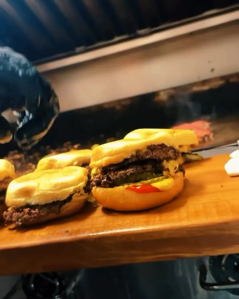 On the line at Better Burgers: finished cheeseburgers on soft potato buns lined up on the wooden board beside the flat-top, a gloved hand at work behind them.