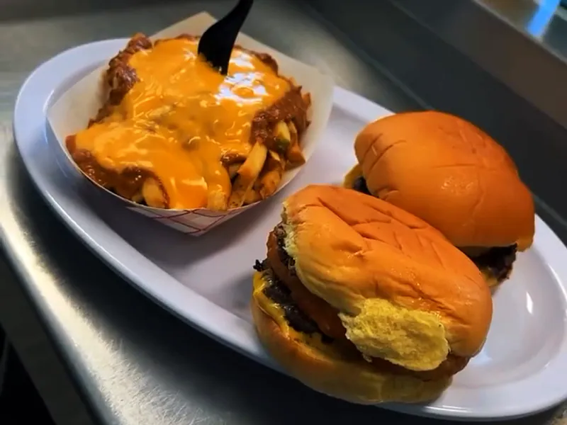 An oval diner plate on the counter with two cheeseburgers and a paper boat of fries smothered in cheese sauce and chili.