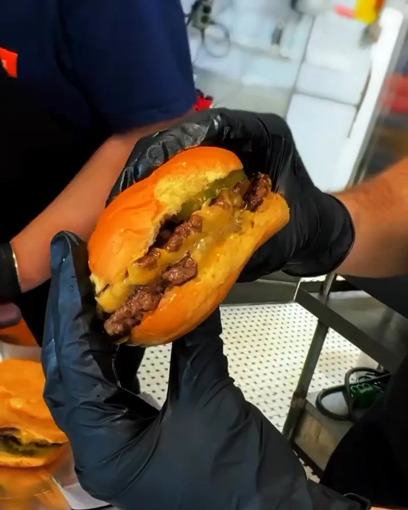 Gloved hands pull a cheeseburger open to show smashed patties, melted American cheese and pickles.
