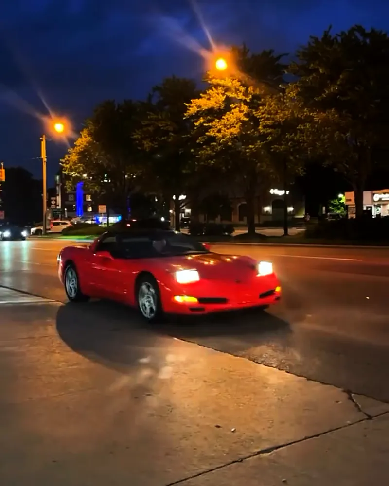 A red classic Corvette cruises past on Woodward at night, headlights on, under the orange street lamps.