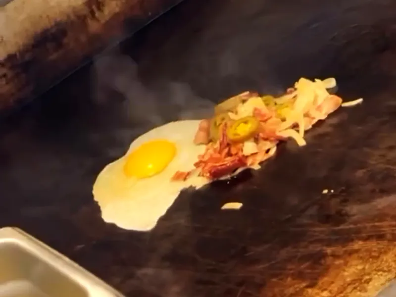 An egg frying sunny-side up on the flat-top beside chopped toppings.