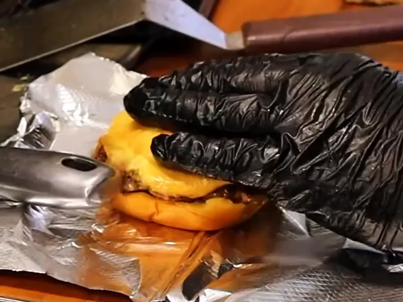 A gloved hand presses a double cheeseburger onto a sheet of foil, ready to wrap.