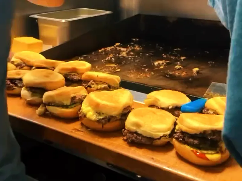 A long row of finished cheeseburgers on soft buns lined up on the wooden board beside the flat-top.