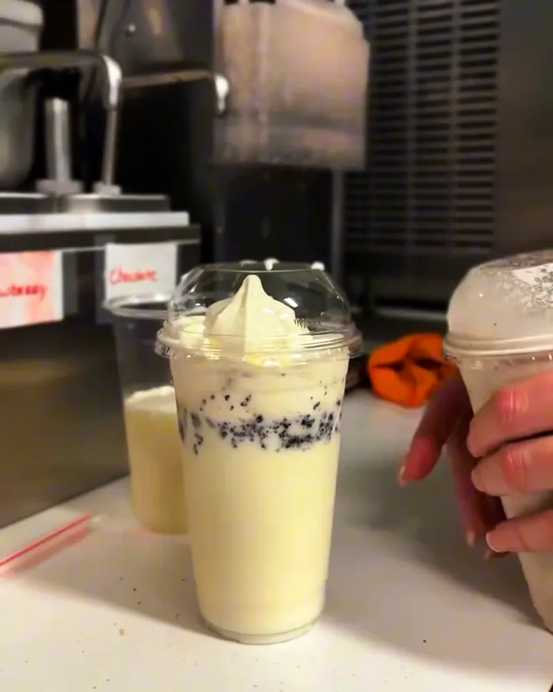 Milkshakes in domed cups at the shake machine; the front one is cookies-and-cream-style.
