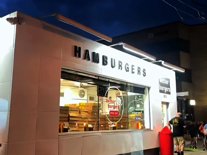 The white Better Burgers building lit up at blue hour, the big front window glowing and a few people by the door.