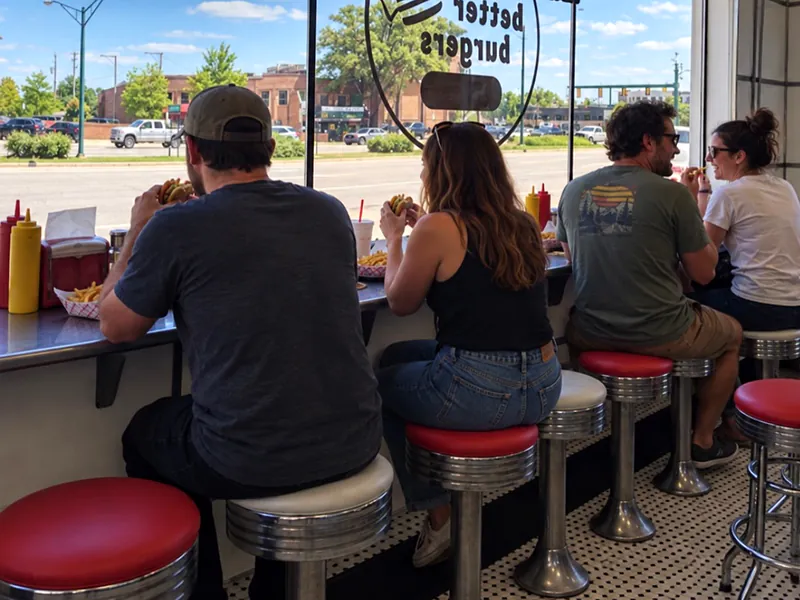 12:30 PM. Lunchtime customers on red-and-chrome stools at the front window counter, the Better Burgers decal on the glass and Woodward outside.