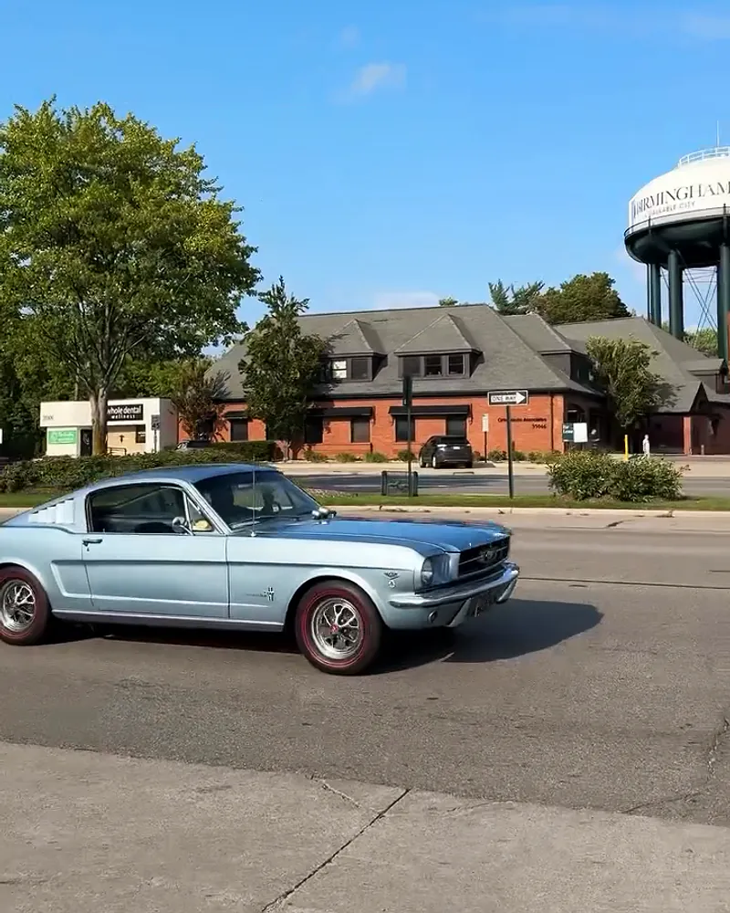 A silver-blue classic Mustang fastback rolls down Woodward in the sun, the Birmingham water tower at the edge of the frame.
