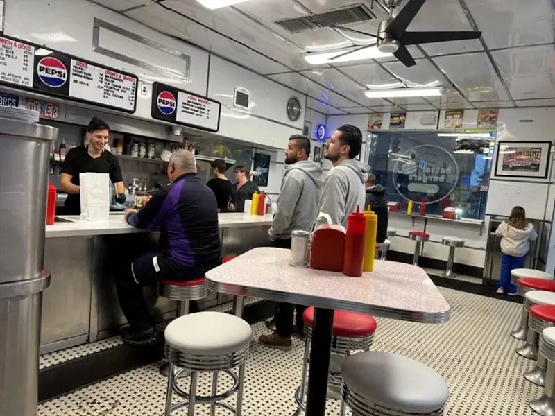 Inside Better Burgers: a white-and-chrome diner counter with red and white stools, menu boards overhead, a cook bagging a takeout order and customers on a black-and-white penny-tile floor.