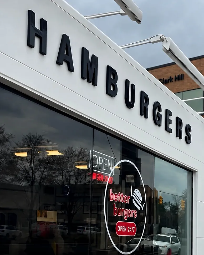 The white Better Burgers storefront by day: big black HAMBURGERS letters, a lit OPEN sign and the red round Better Burgers OPEN 24/7 decal in the window.